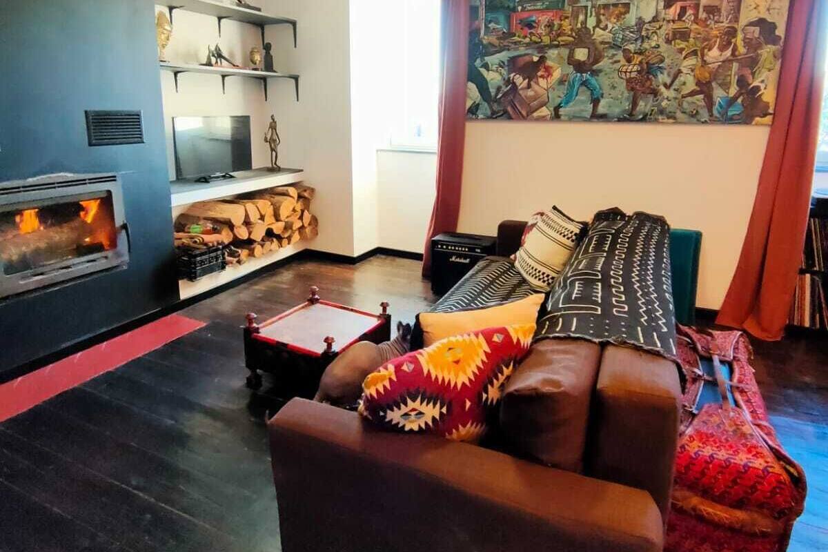 Uma sala de estar com lareira, ventilador de teto, sofá com almofadas estampadas e um mural colorido na parede.