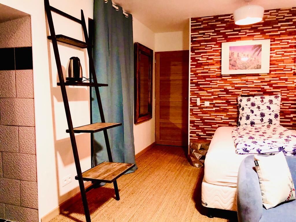 A compact room with a bed, a wooden ladder shelf, a framed picture on the wall, and a patterned pillow.