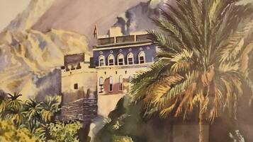Un edificio tradicional con ventanas de arco y techo plano, rodeado de palmeras y montañas.