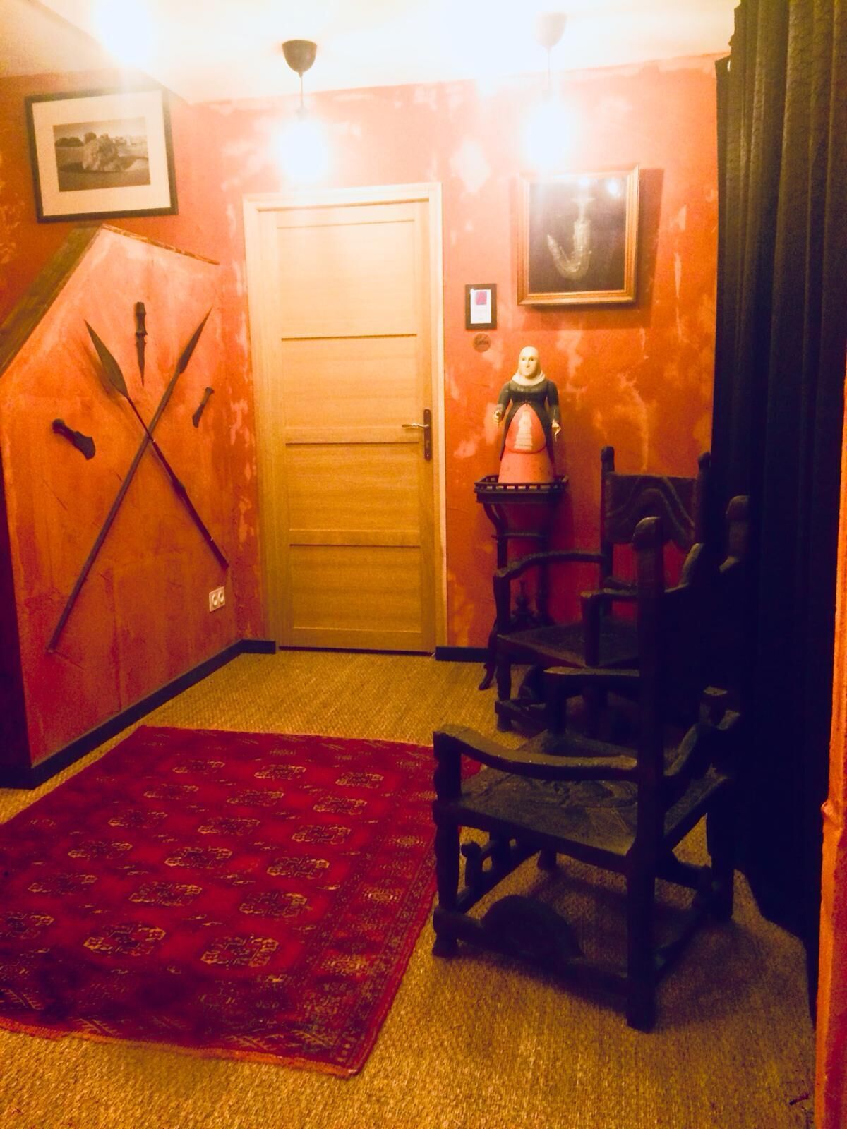 Una habitación con puerta de madera, alfombra roja, una silla de madera y una lámpara fijada a la pared.