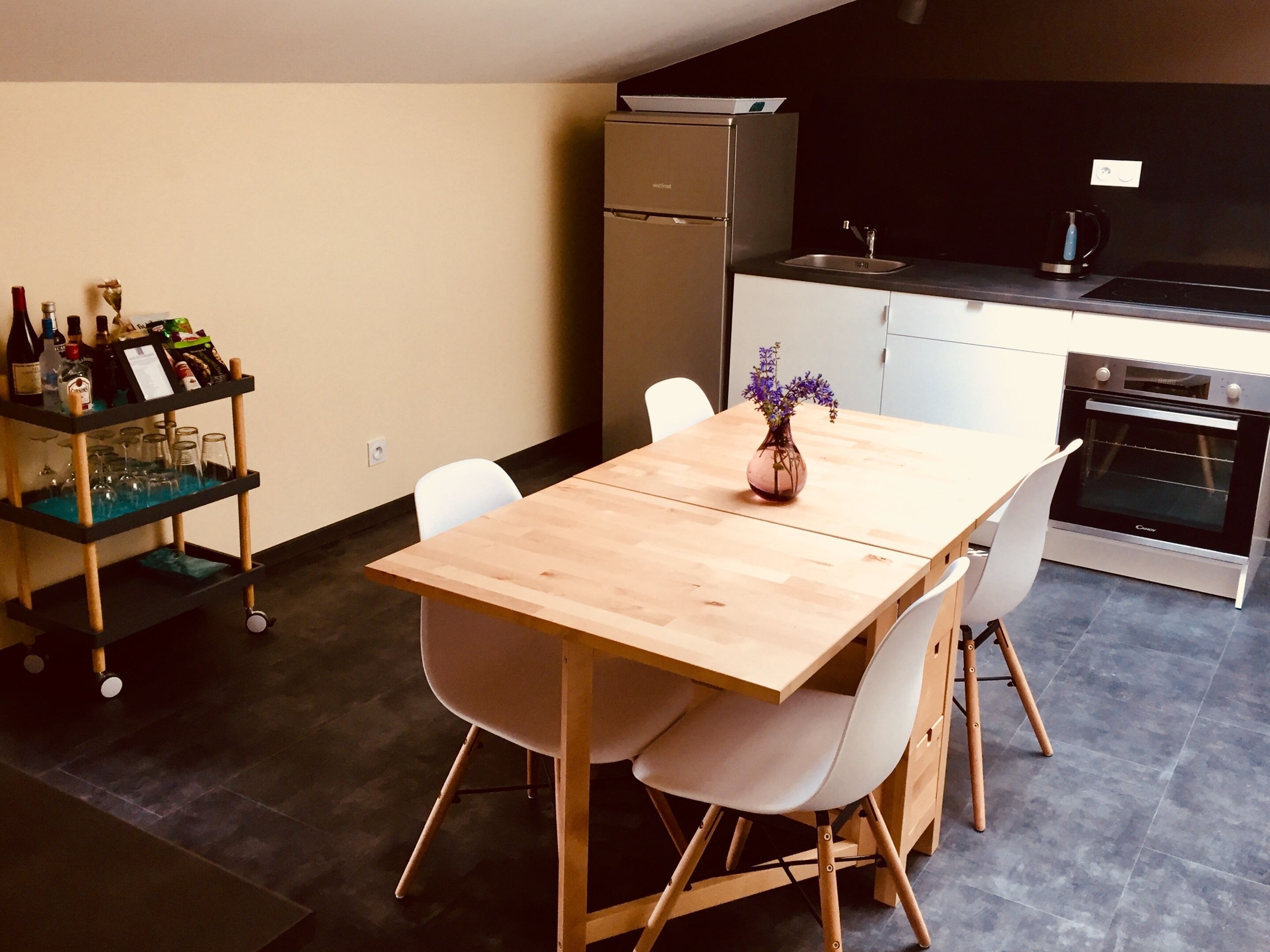 Une cuisine moderne avec une table et des chaises en bois, un réfrigérateur, une machine à café et un meuble à vin contenant des bouteilles.