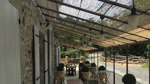 Un patio al aire libre con mesas y sillas, una zona techada con techo de paja, y vista a una carretera con troncos.