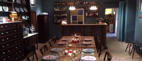 A dining area with a long wooden table set for six, surrounded by chairs. There are shelves with various items on the left side, and a bar area with bottles in the background.