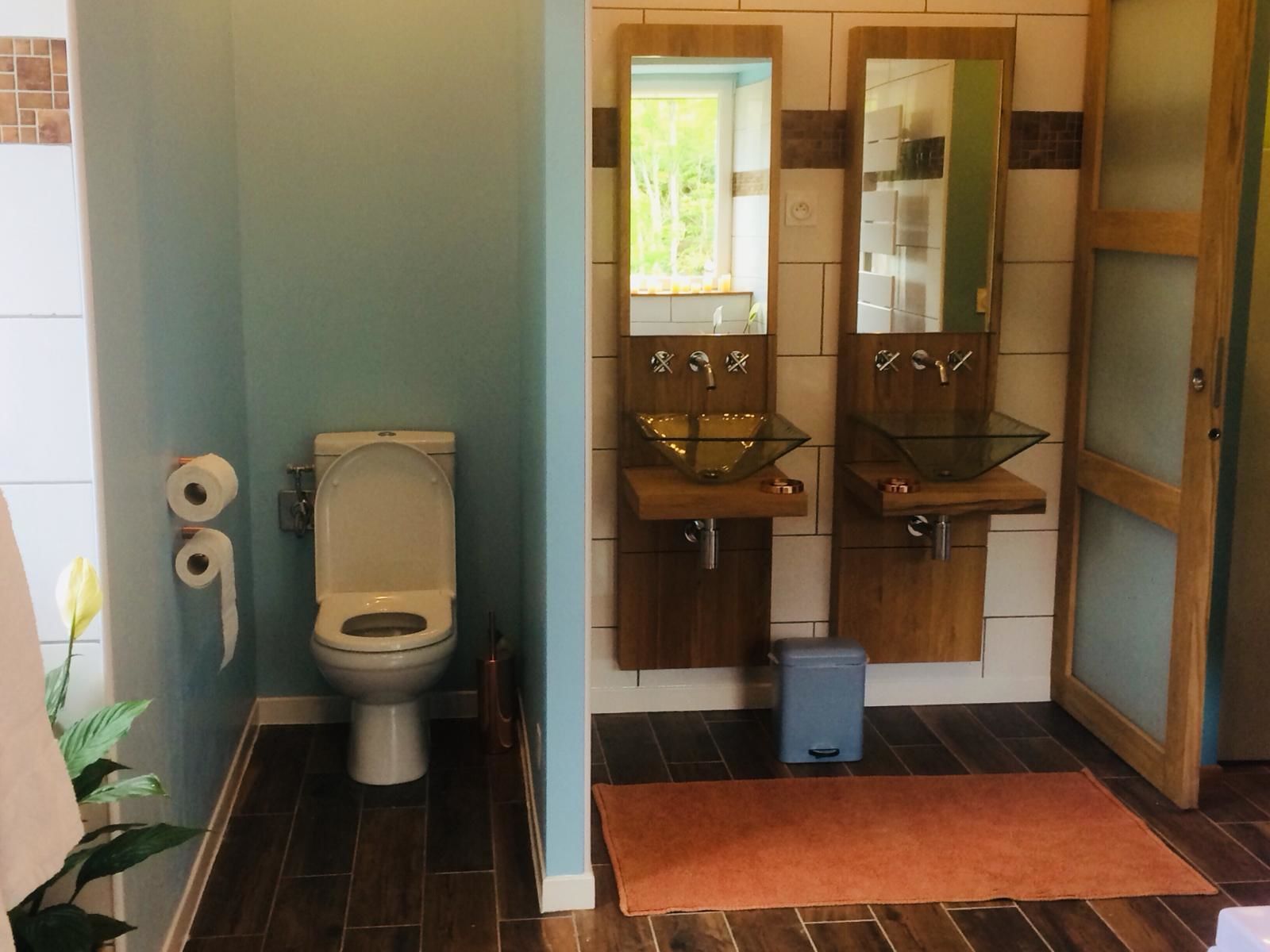 A bathroom with a toilet, two sinks with glass bowls, a trash bin, and a brown rug on the floor.