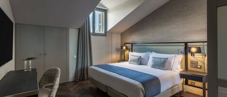 A modern hotel room with a large bed, a desk, and a chair. There is a window with curtains, a lamp, and a nightstand with a phone.