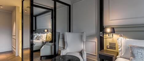 A hotel room with a mirrored closet, a bed with white linens, a white armchair, a small round table, and a bedside lamp.