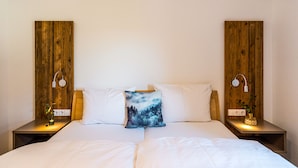 Een slaapkamer met een houten hoofdbord, witte beddengoed, een decoratief kussen, bedlampen en een kleine plant op het nachtkastje.