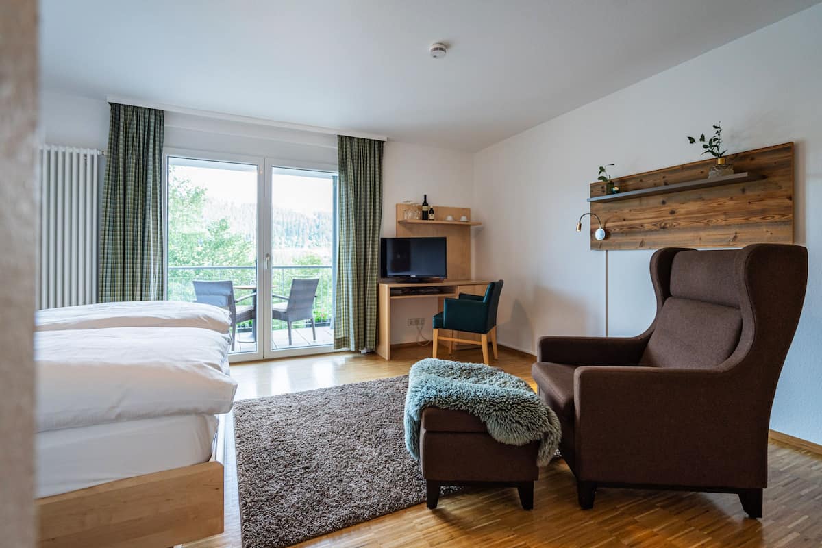 A modern hotel room with a bed, a desk with a computer, a TV, a brown armchair, and a balcony with a view.