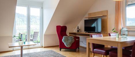 Una sala de estar moderna con televisor, una zona de comedor con mesa y sillas, un sillón rojo y un ventanal con vistas a árboles.