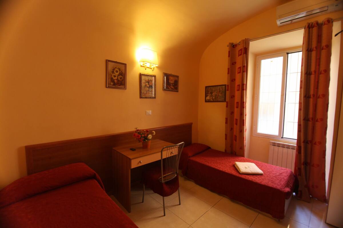 A small, cozy room with two single beds, a desk with a chair, a wall-mounted lamp, and a window with curtains.