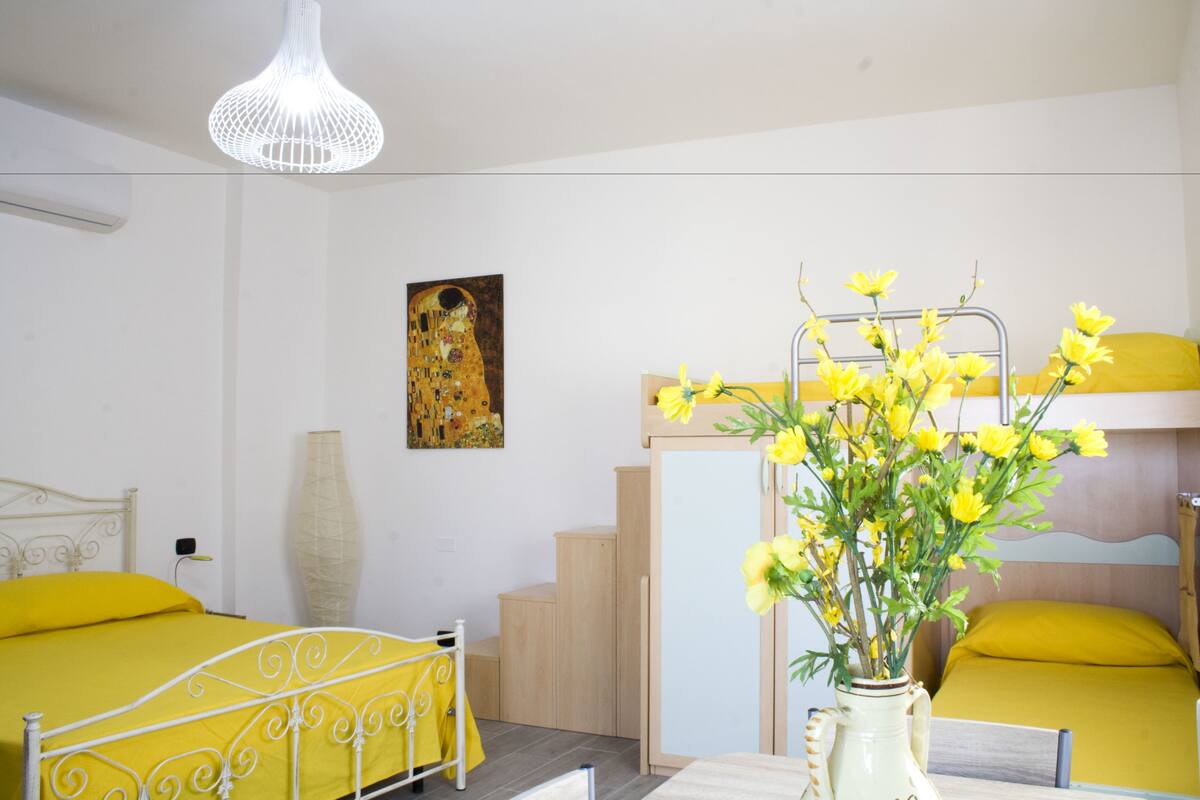 Una habitación con literas, una mesita de noche con un florero, un cuadro en la pared y una lámpara colgante.