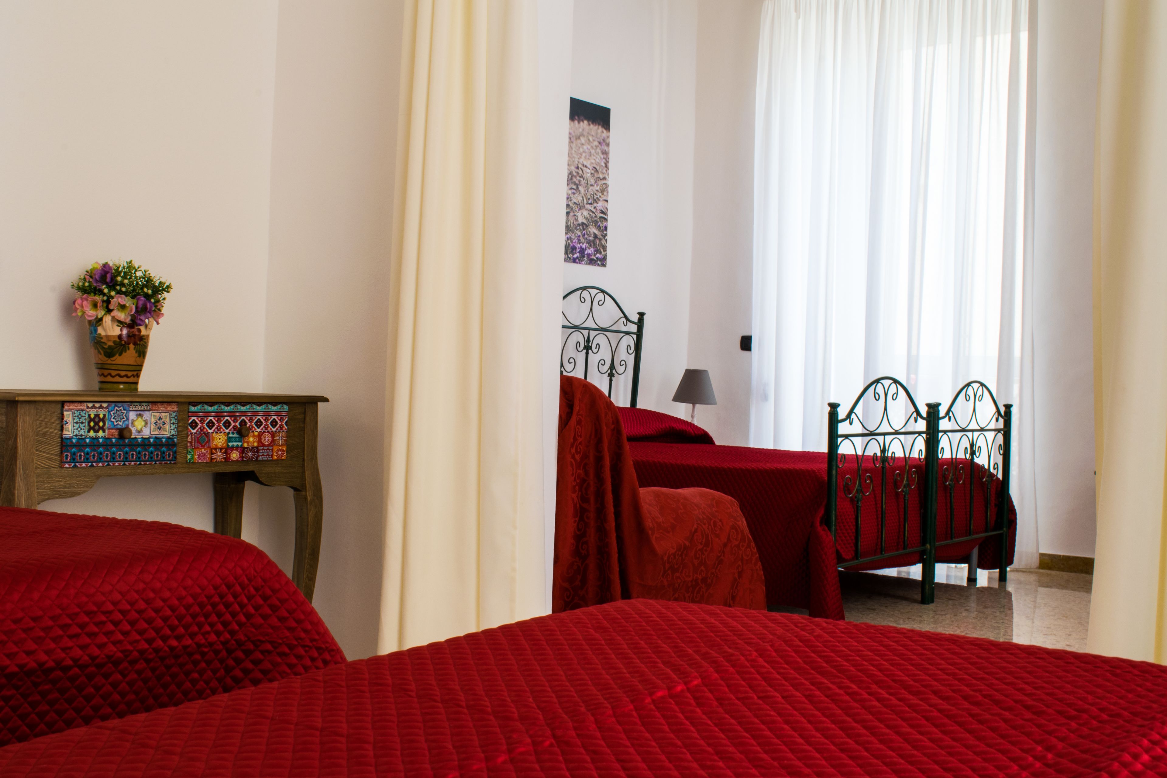 A bedroom with two beds, a wooden nightstand with a decorative vase, and a view of a garden through sheer curtains.