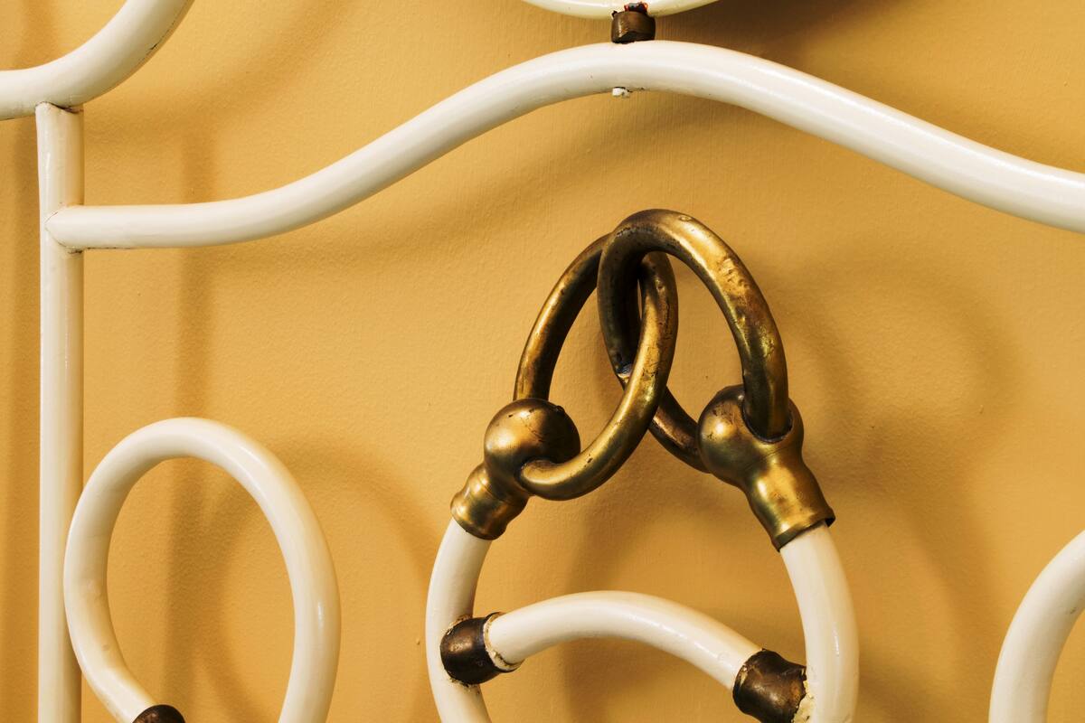 Primer plano de una estructura de cama metálica blanca con detalles de latón sobre un muro amarillo.