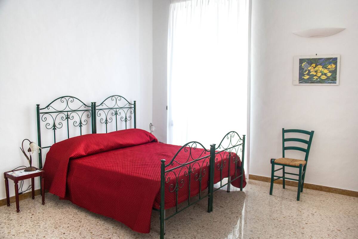 Una habitación con estructura metálica para la cama, una colcha roja, una mesita de noche con una lámpara, una silla y un cuadro en la pared.