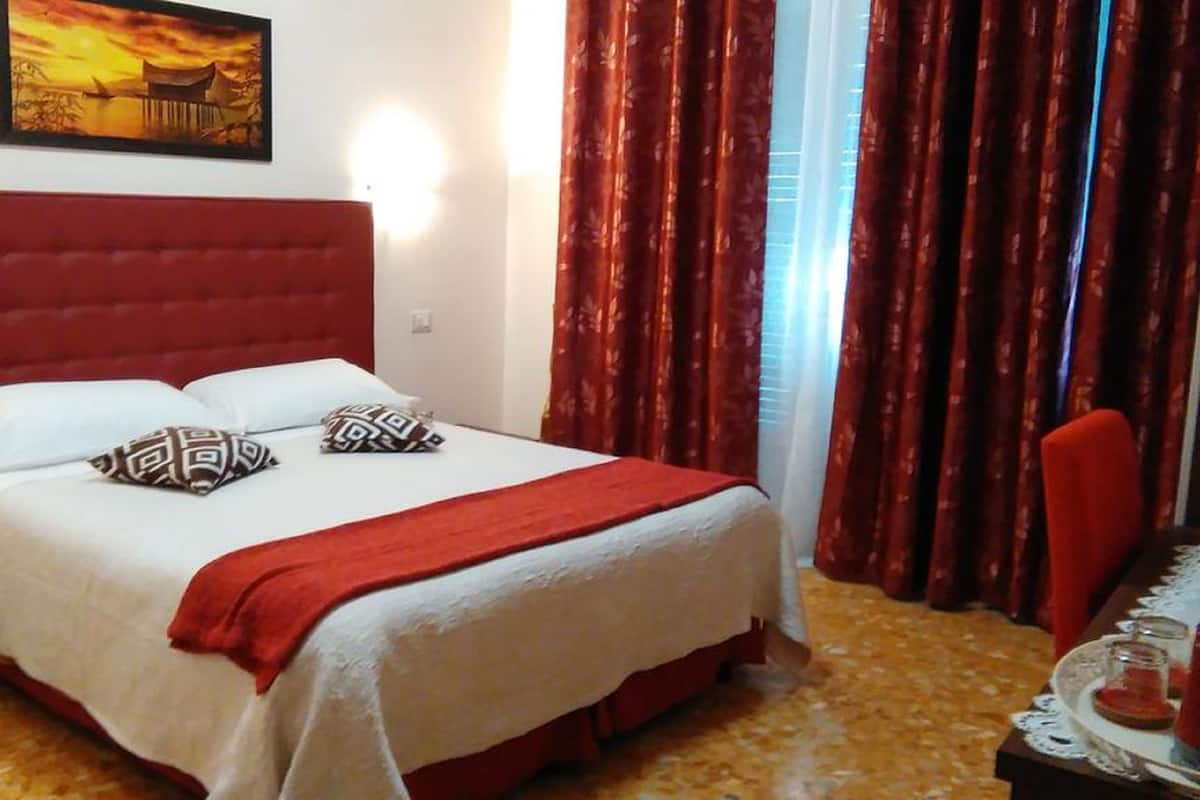 Ein Hotelzimmer mit einem roten, gepolsterten Bett, weißer Bettwäsche, gemusterten Kissen, einer roten Tagesdecke, Holzboden, einem Gemälde über dem Bett und roten Vorhängen.