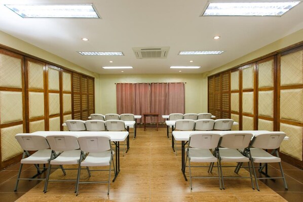 Un salón de conferencias con sillas y mesas blancas dispuestas en filas, un tabique de madera y un ventanal con cortinas.