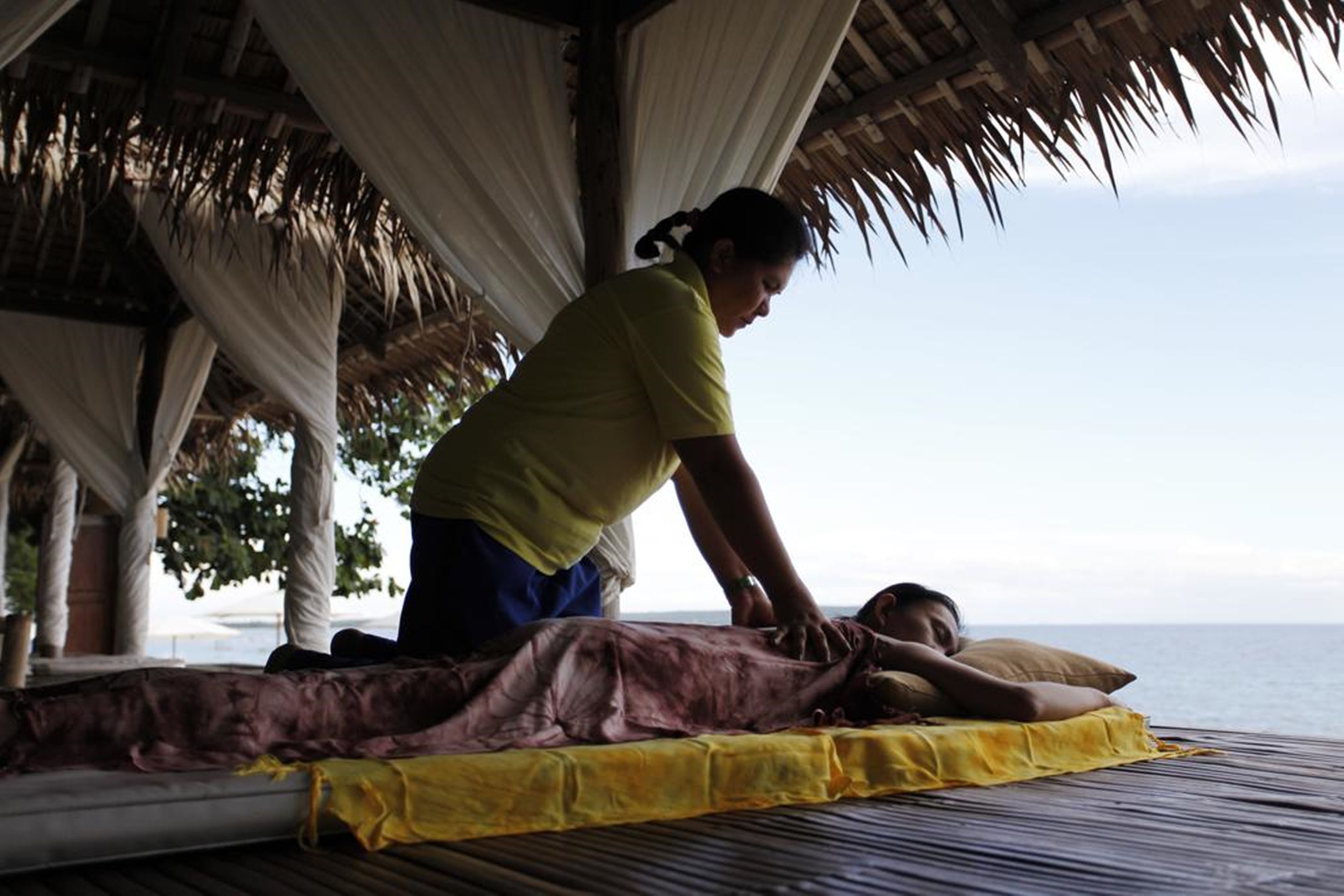 A massage therapy session taking place on a bed under a thatched roof structure with a view of the sea.