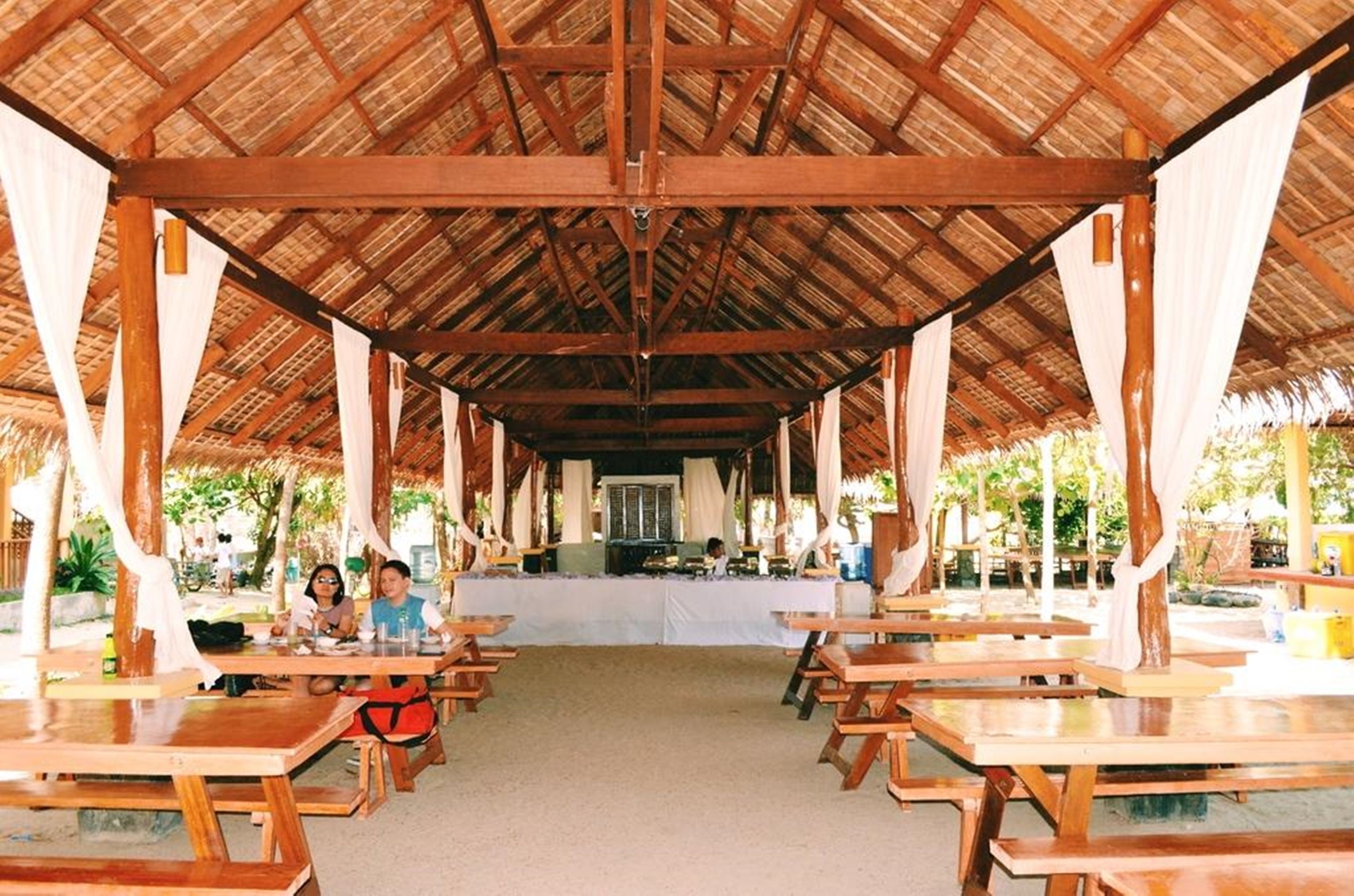 Un área de comedor al aire libre techada, con mesas y bancos de madera, un pasillo central y una estructura de techo de paja.