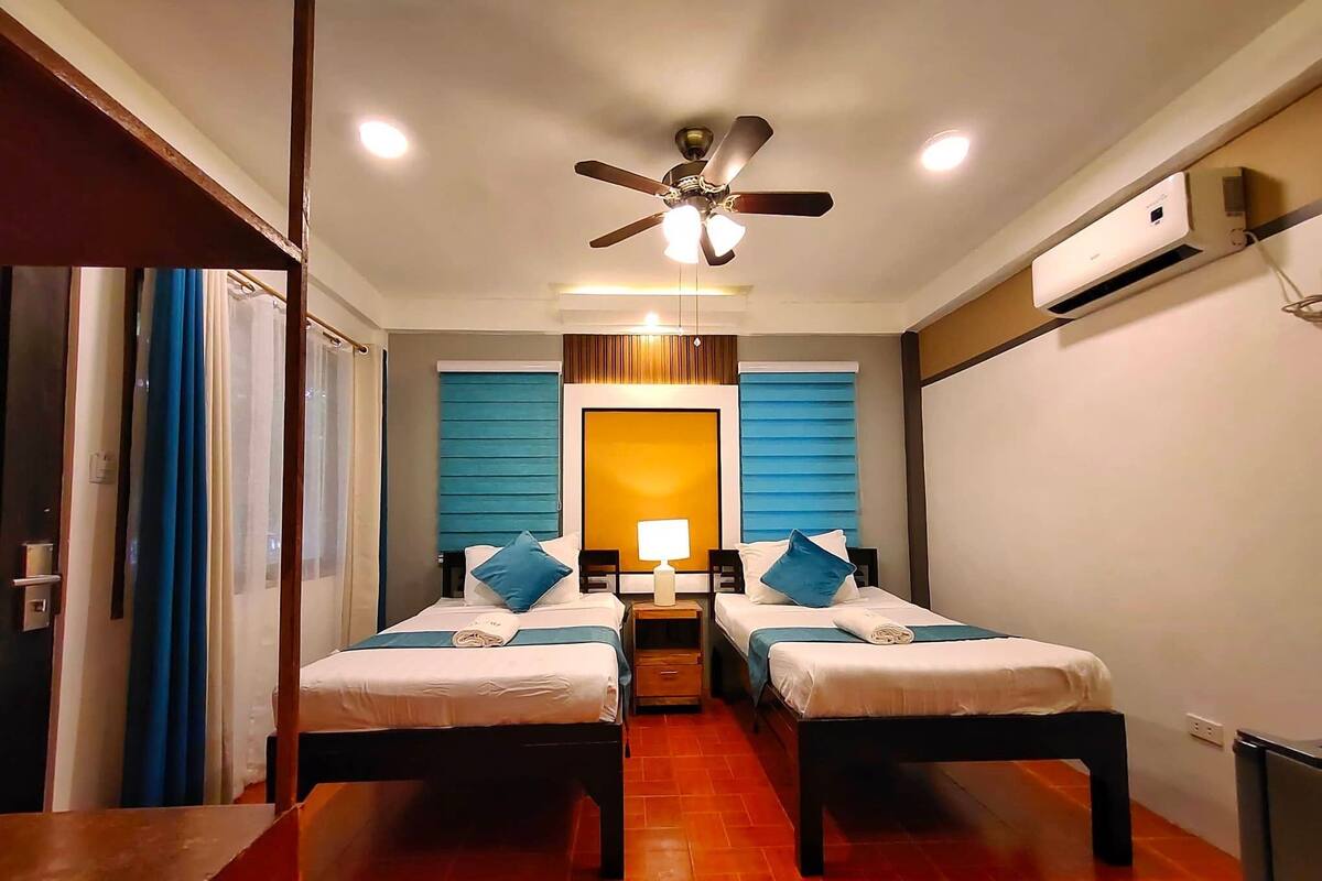Una habitación de hotel con dos camas, ventilador de techo, unidad de aire acondicionado y piso de madera.
