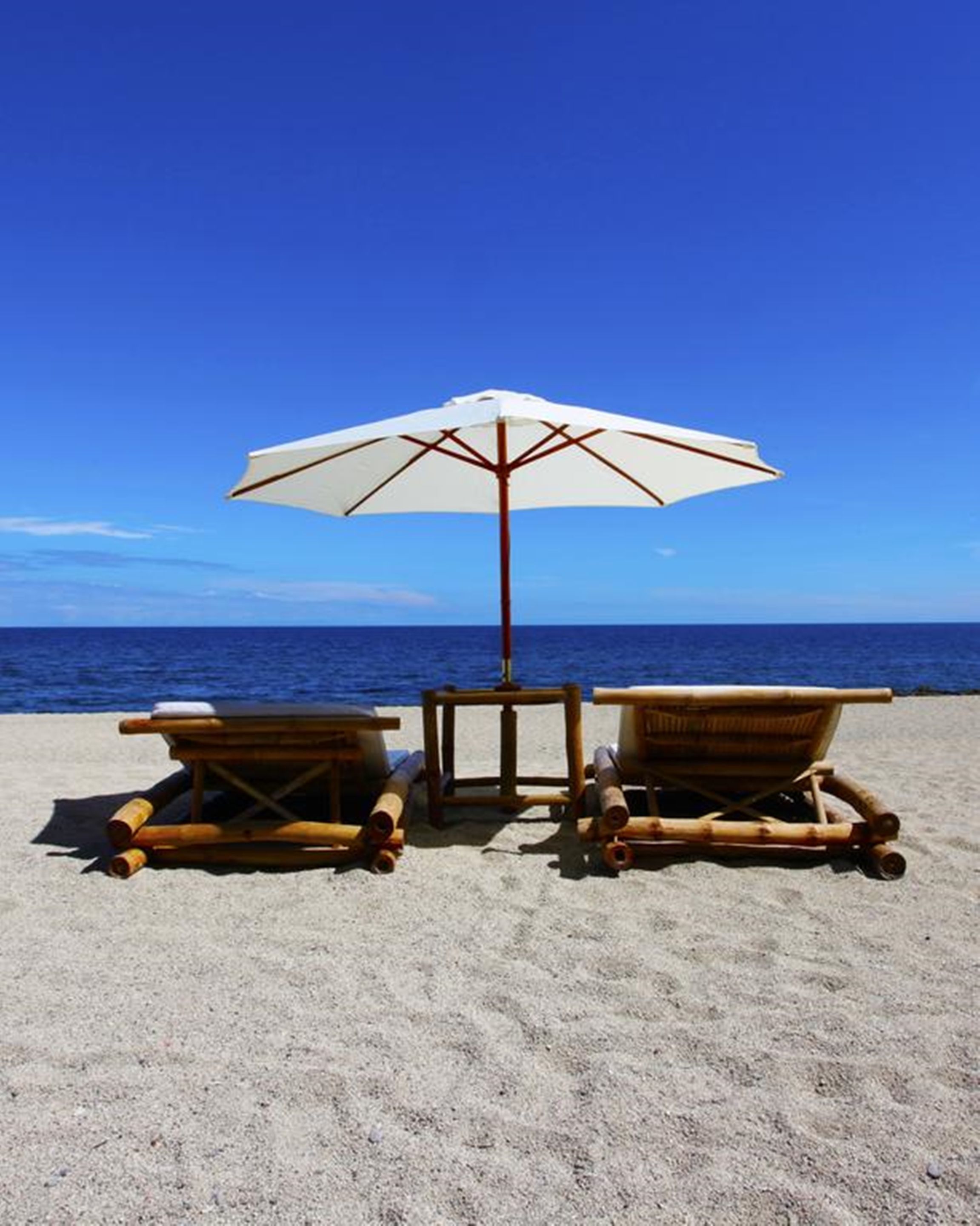 Deux fauteuils de plage en bois, avec un parasol blanc, sur une plage de sable.