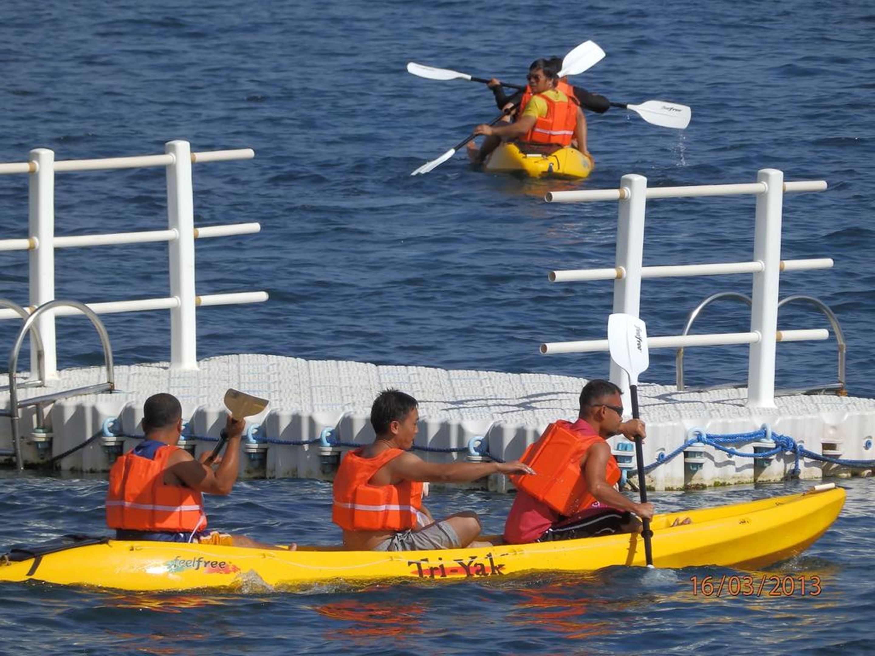 Three people in a yellow kayak with 'feelfree' and 'Tri-Yak' logos, wearing orange life jackets, paddling near a dock with white railings.