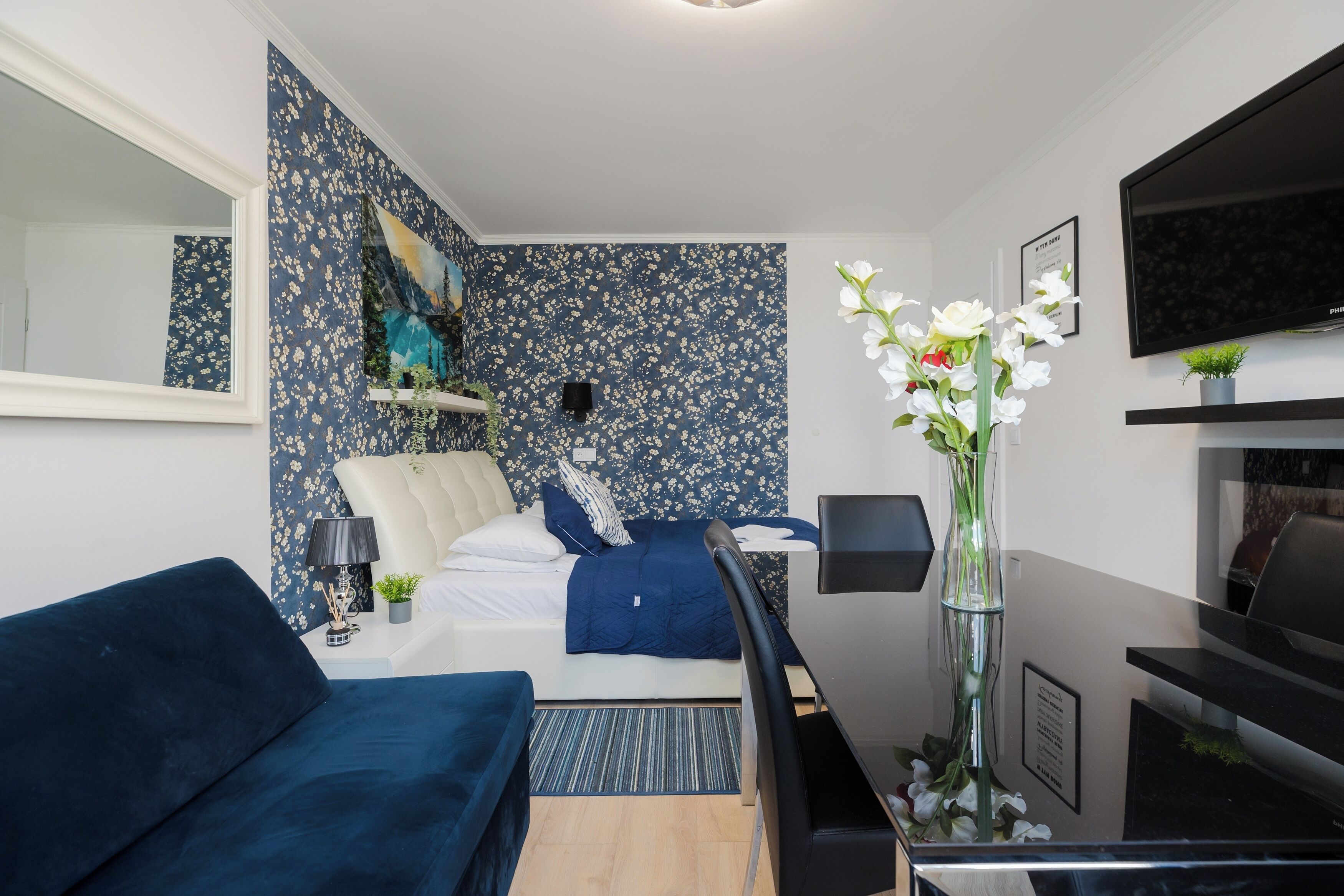 Une chambre d’hôtel moderne avec un lit, un canapé bleu, une télévision et une petite table sur laquelle est posé un vase de fleurs.