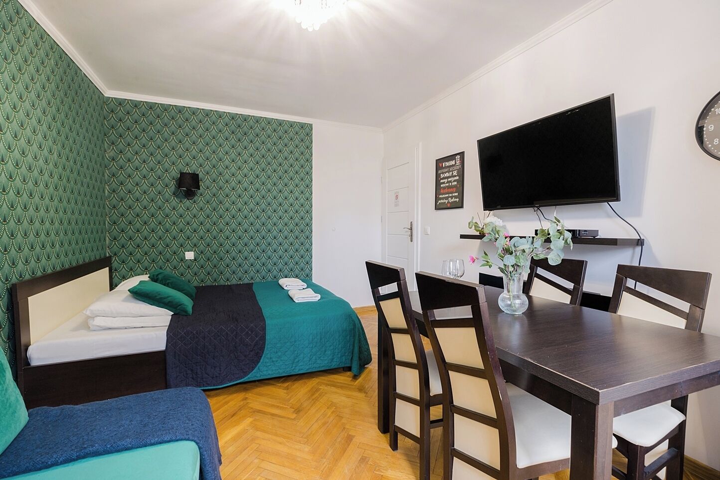 A room with two beds, a dining table with chairs, a flat-screen TV, and a wall-mounted clock.