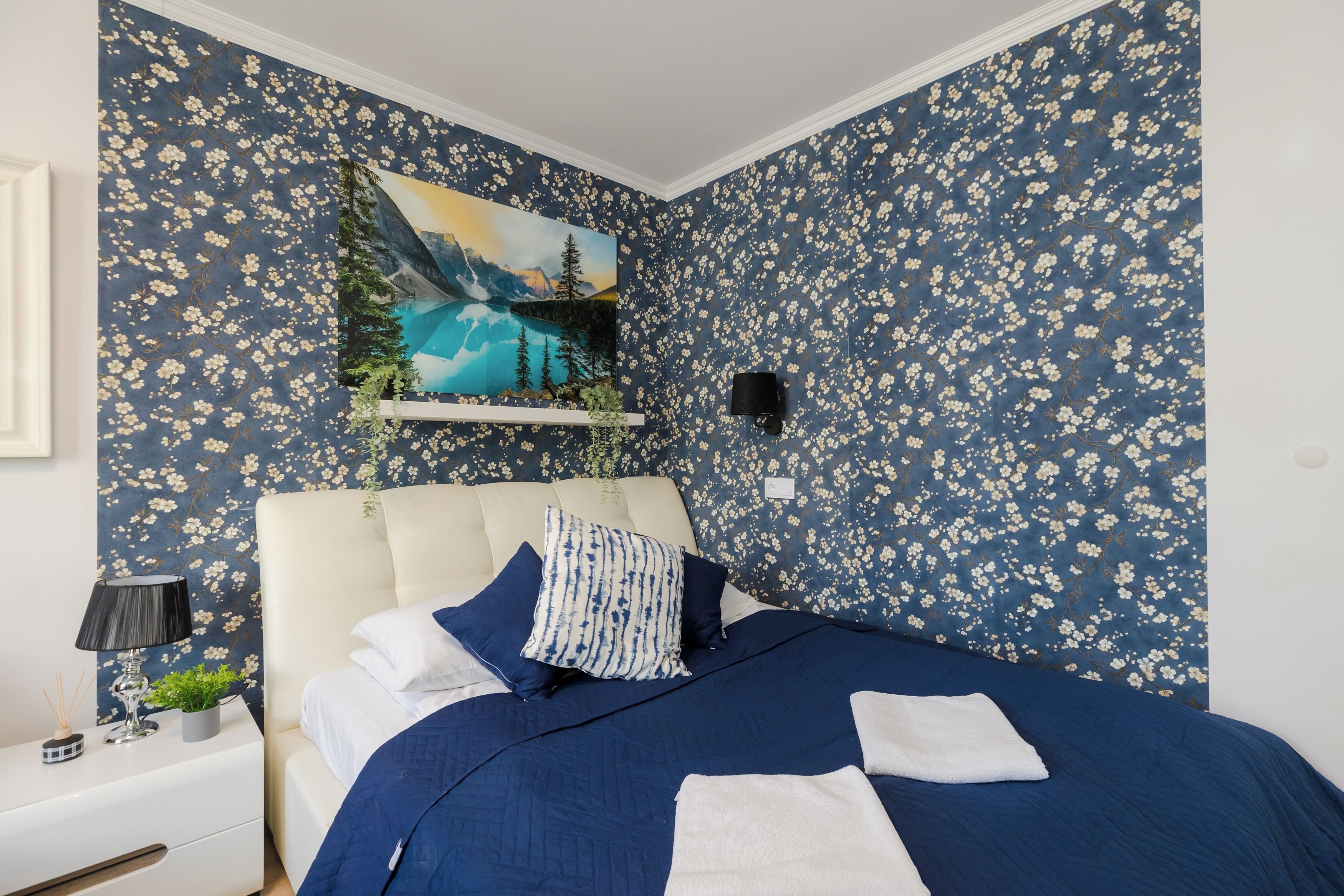 Une chambre à coucher avec un papier peint à motifs floraux, un lit avec une literie bleue, une table de chevet avec une lampe et un tableau au-dessus du lit.