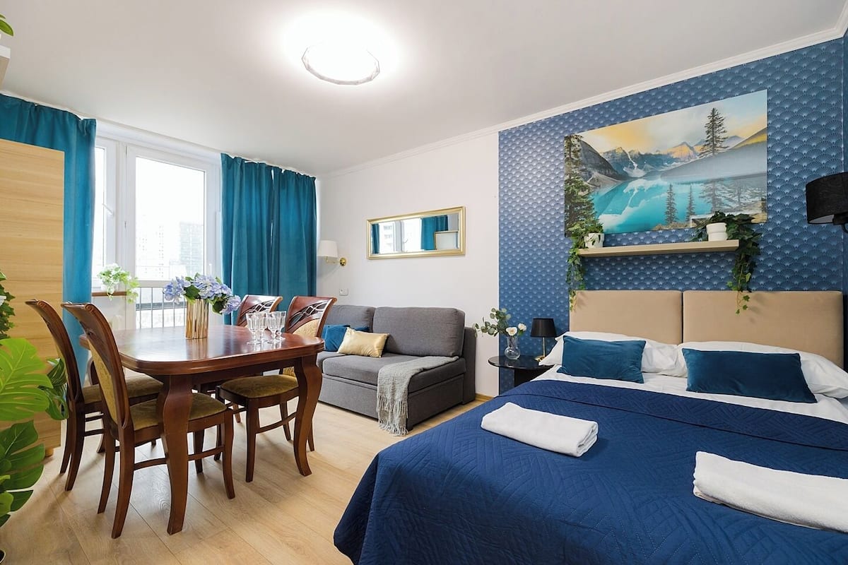A hotel room with a dining table, a sofa, a bed with blue bedding, and a painting of a mountain landscape.