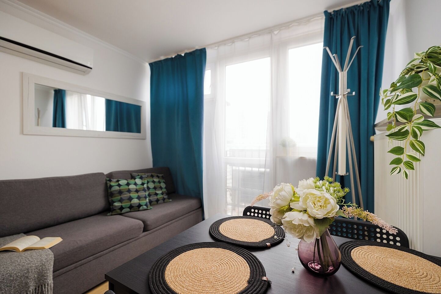 A modern living room with a grey sofa, a coffee table with woven placemats, a vase with white flowers, and a large window with blue curtains.