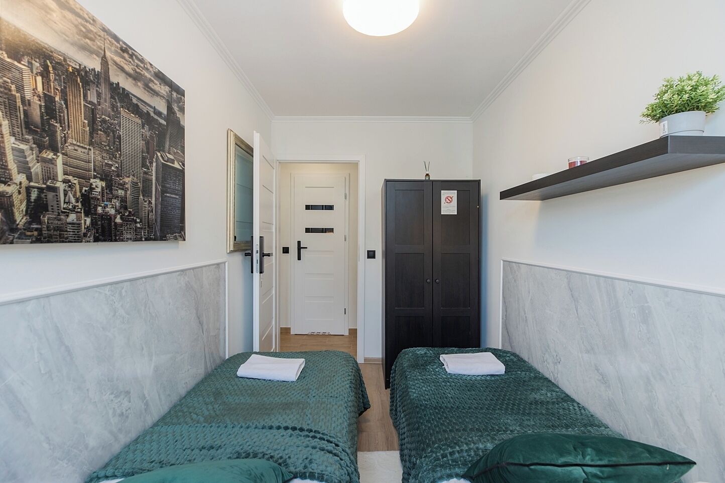 A narrow room with two single beds, a door, a wardrobe, and a cityscape painting on the wall.