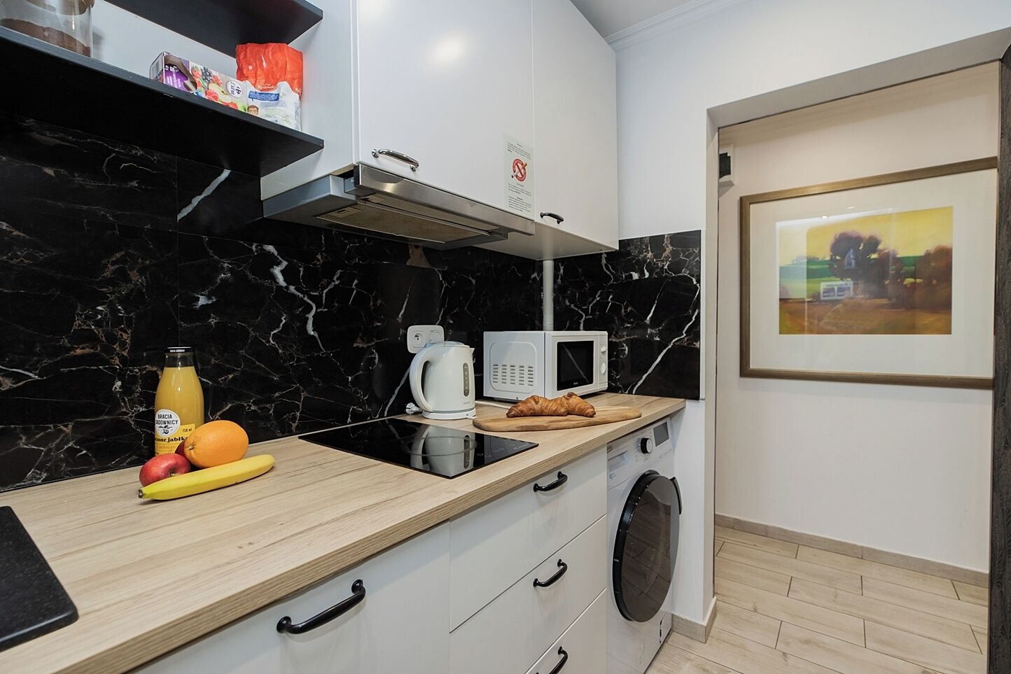 A modern kitchen with a black marble backsplash, white cabinets, a microwave, a kettle, and a wooden countertop with fruits and a croissant.