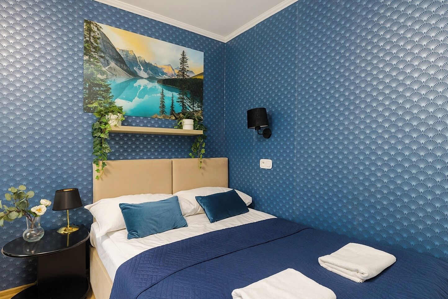 A bedroom with a bed, a nightstand, a wall-mounted lamp, and a painting of a mountain landscape.