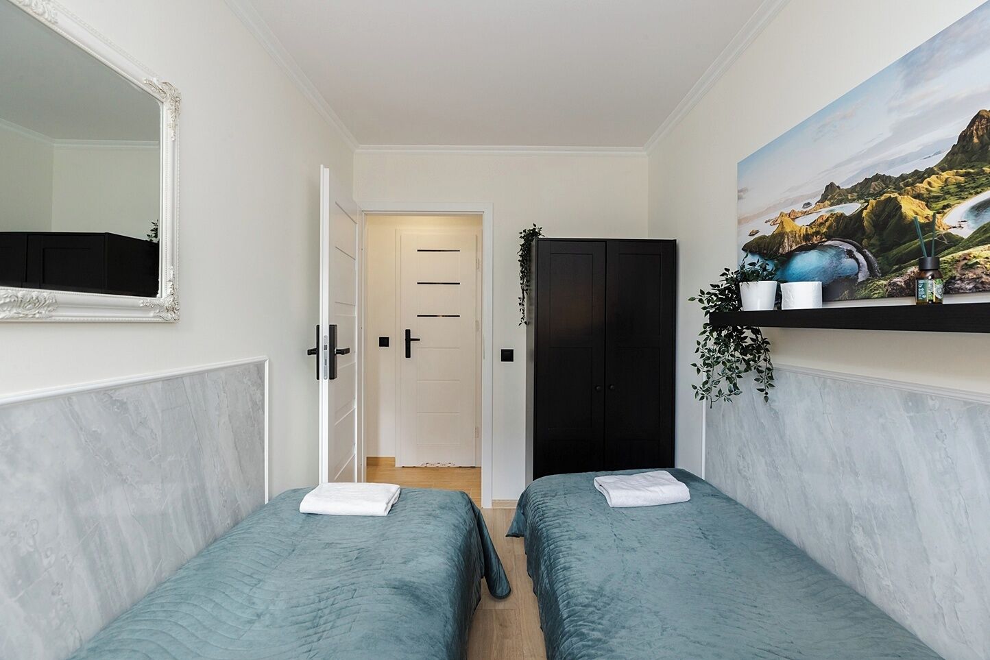 A room with two beds, a large mirror, a door leading to another room, and a shelf with plants and a painting.