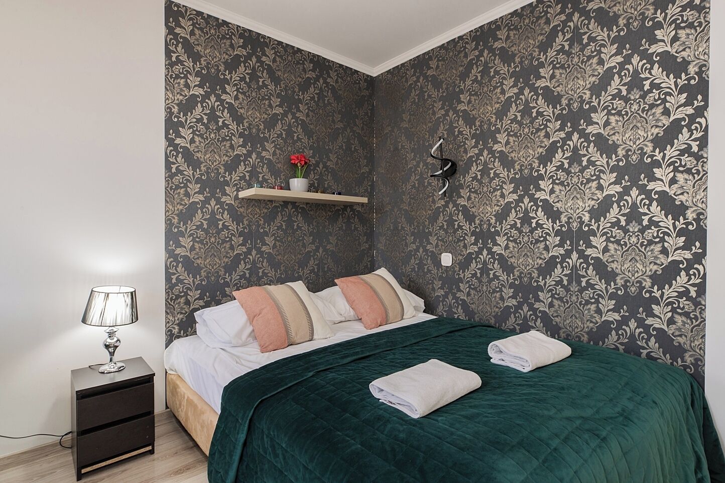 A bedroom with a bed, a nightstand, a lamp, a shelf with a vase, and a wall with a patterned wallpaper.