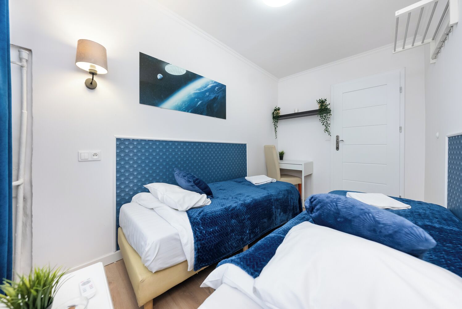 Un dormitorio con dos camas, una lámpara de pared, un cuadro de espacio y un escritorio pequeño con una planta.