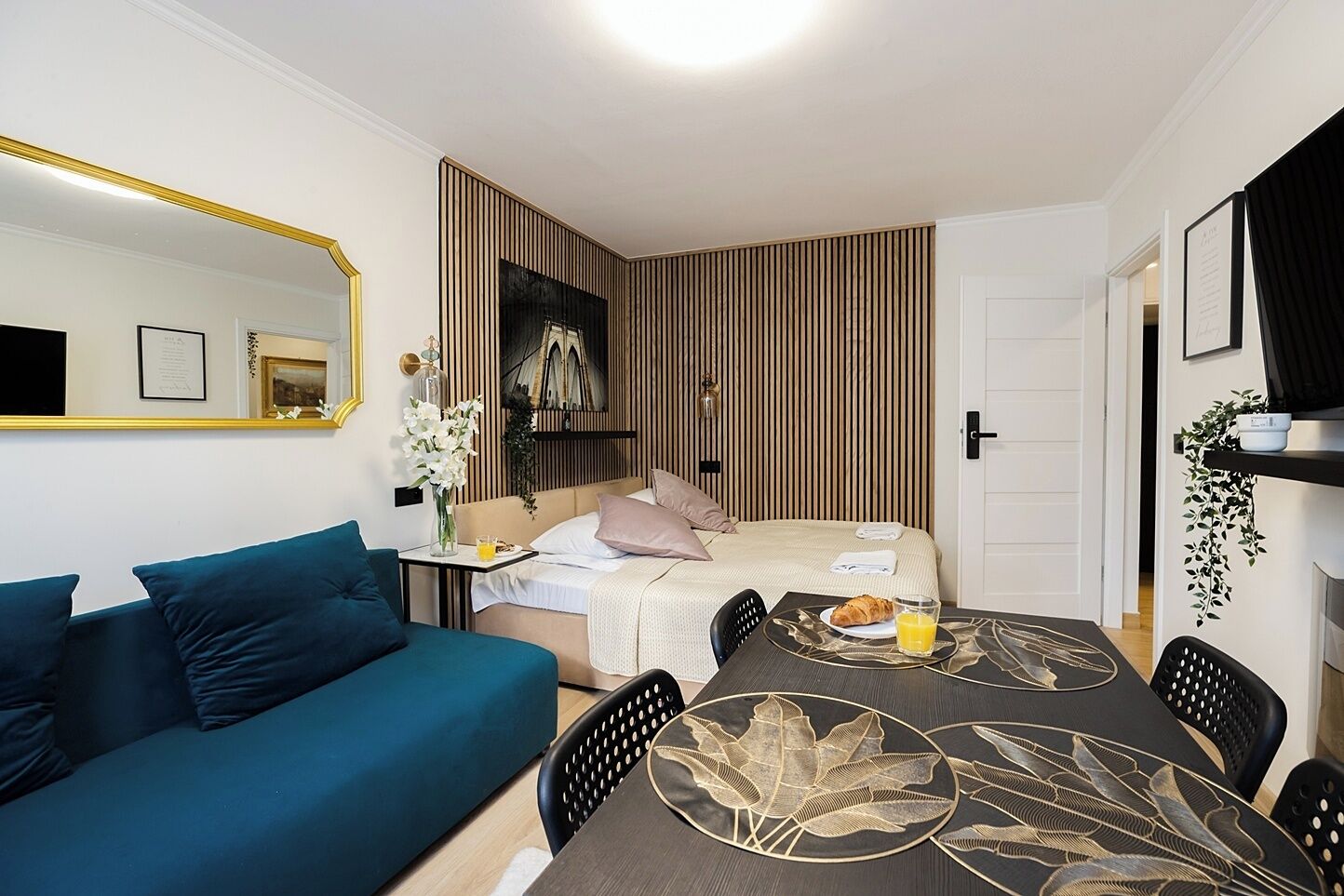 A hotel room with a bed, a sofa, a dining table with a decorative table runner, a mirror, and a plant.