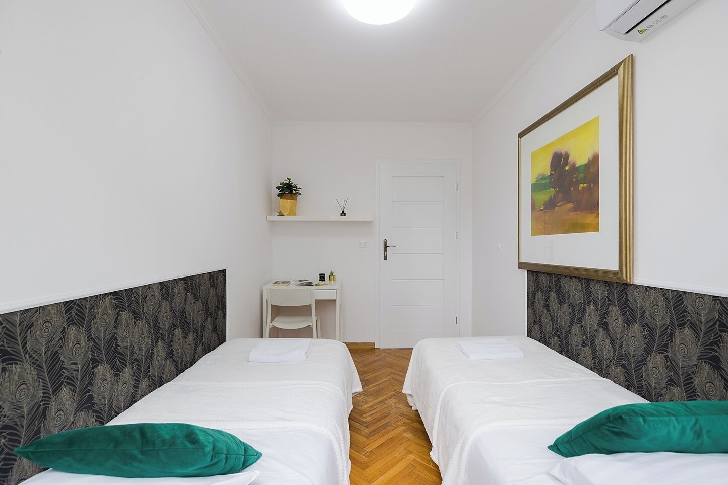 A room with two single beds, a desk, a chair, a shelf with a plant, and a framed picture on the wall.