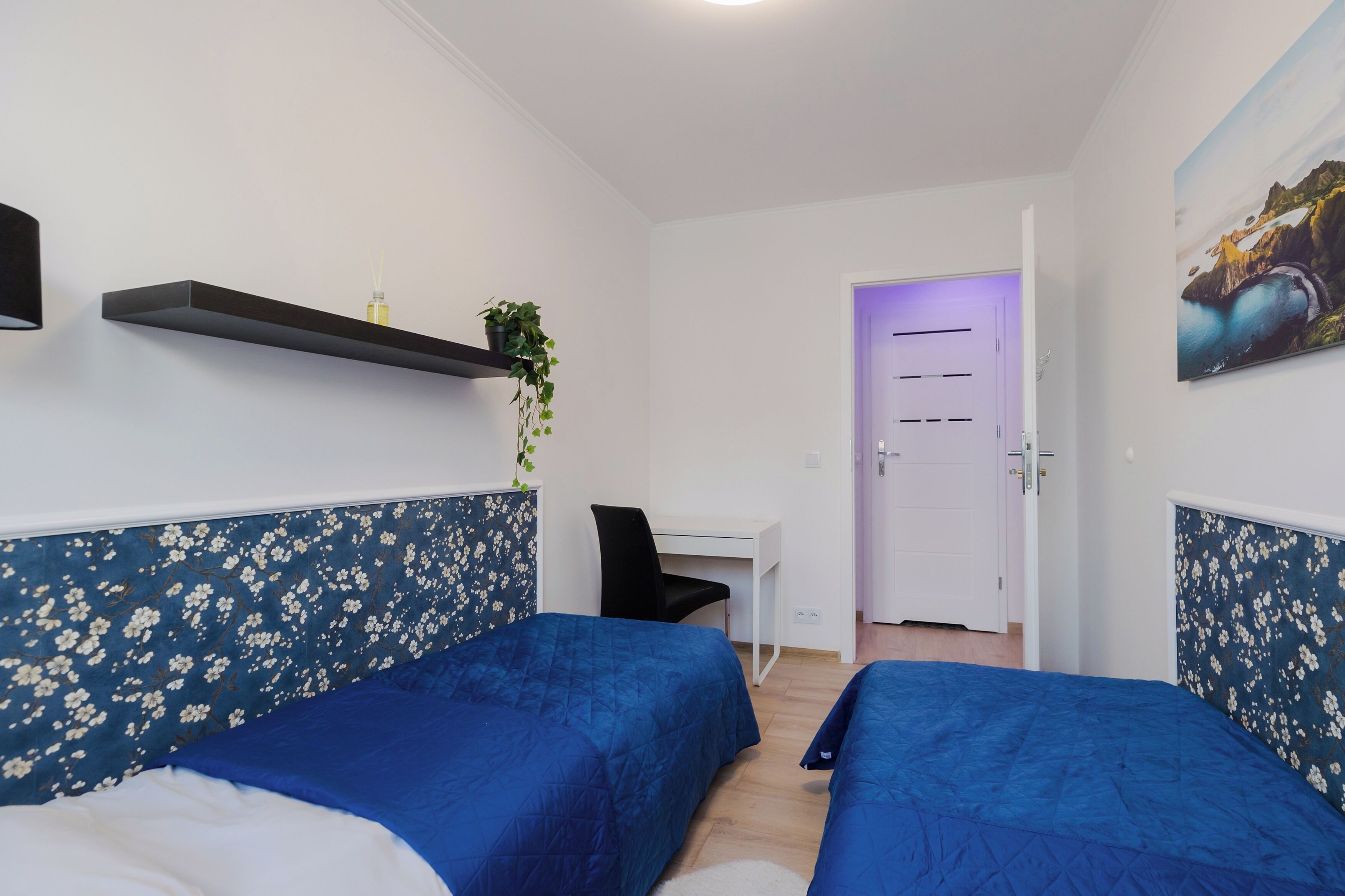 Habitació amb dos llits, un escriptori i una cadira. Hi ha una porta amb llum morada i un quadre a la paret.