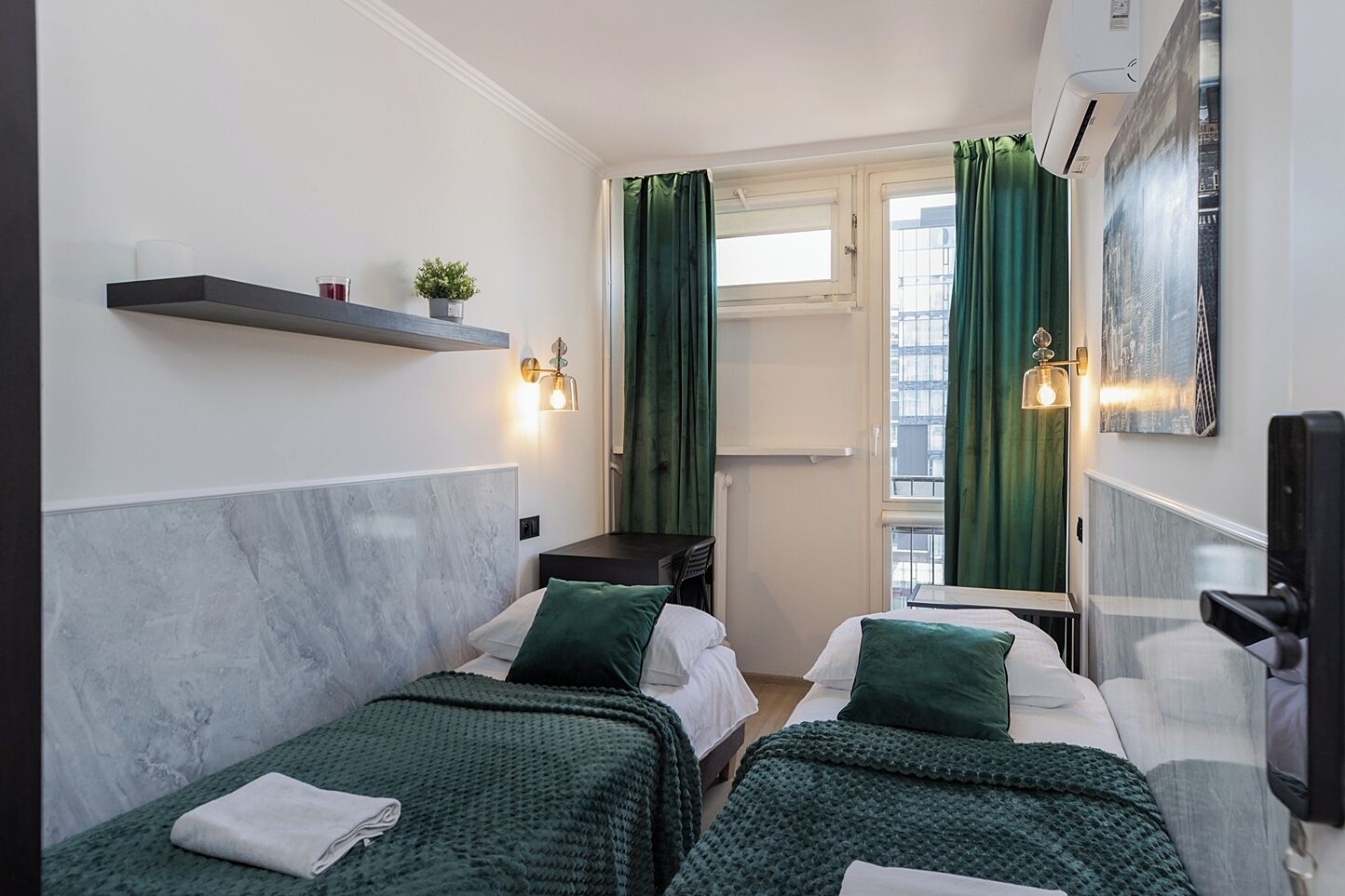 Habitació d'hotel amb dos llits, una taula petita, una estant amb una planta i una finestra amb cortines.