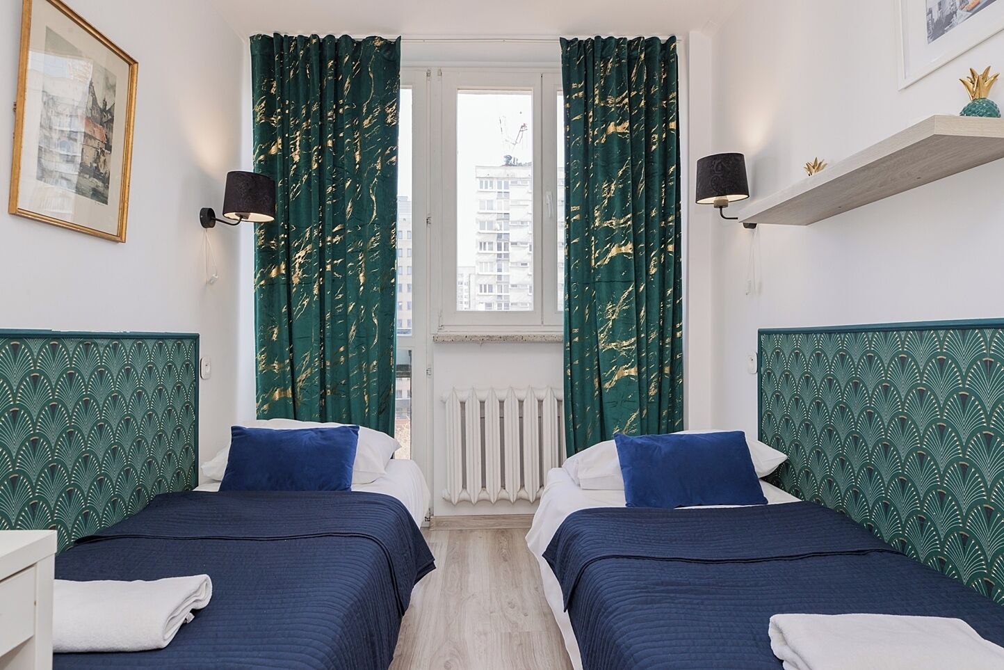 A room with two beds, green patterned headboards, white bedding, a window with curtains, a radiator, and a framed picture on the wall.