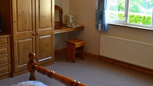 Un dormitorio con un armario de madera, una cama de madera con cabecera, una mesita de madera, un radiador y una ventana con vistas a zonas verdes.