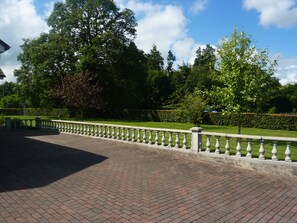 Une allée dallée, bordée d’une balustrade et entourée d’arbustes et d’arbres bien entretenus.