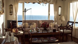 Ein Wohnzimmer mit einer Couch, Sesseln, einem Couchtisch mit Zeitschriften, einem Fernseher und Blick aufs Meer.