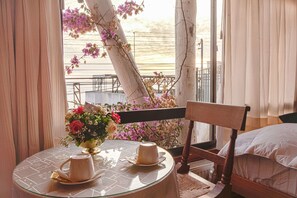 Quarto com mesa de vidro redonda, duas xícaras brancas, um vaso com flores, uma cadeira de madeira e uma cama com lençóis brancos.