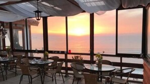 Ein Außenbereich zum Essen mit Tischen und Stühlen, große Fenster mit Blick auf den Sonnenuntergang über dem Meer.