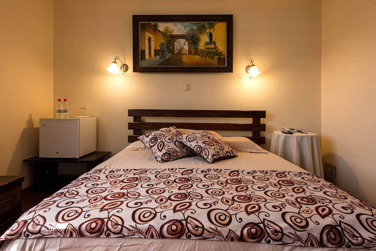 A hotel room with a bed, a painting on the wall, a small table with a lamp, and a minibar.