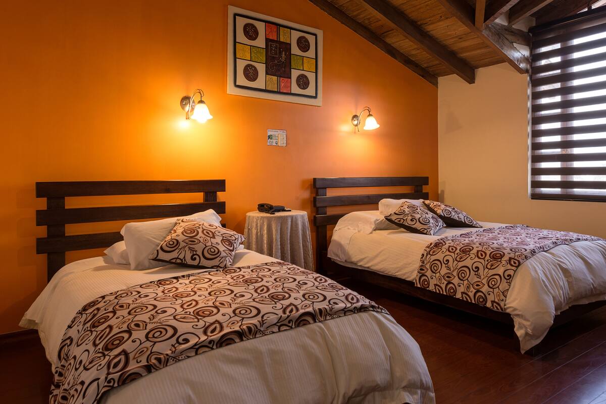 A room with two beds, each with patterned bedding, a small round table, and wall-mounted lights.