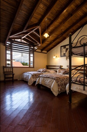 Una habitación con vigas de madera, una cama con ropa de cama estampada, una silla y un ventanal con vistas a un edificio de techo rojo.