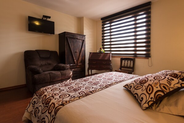 Un dormitorio con cama, una silla, armario, cómoda y televisor en la pared.